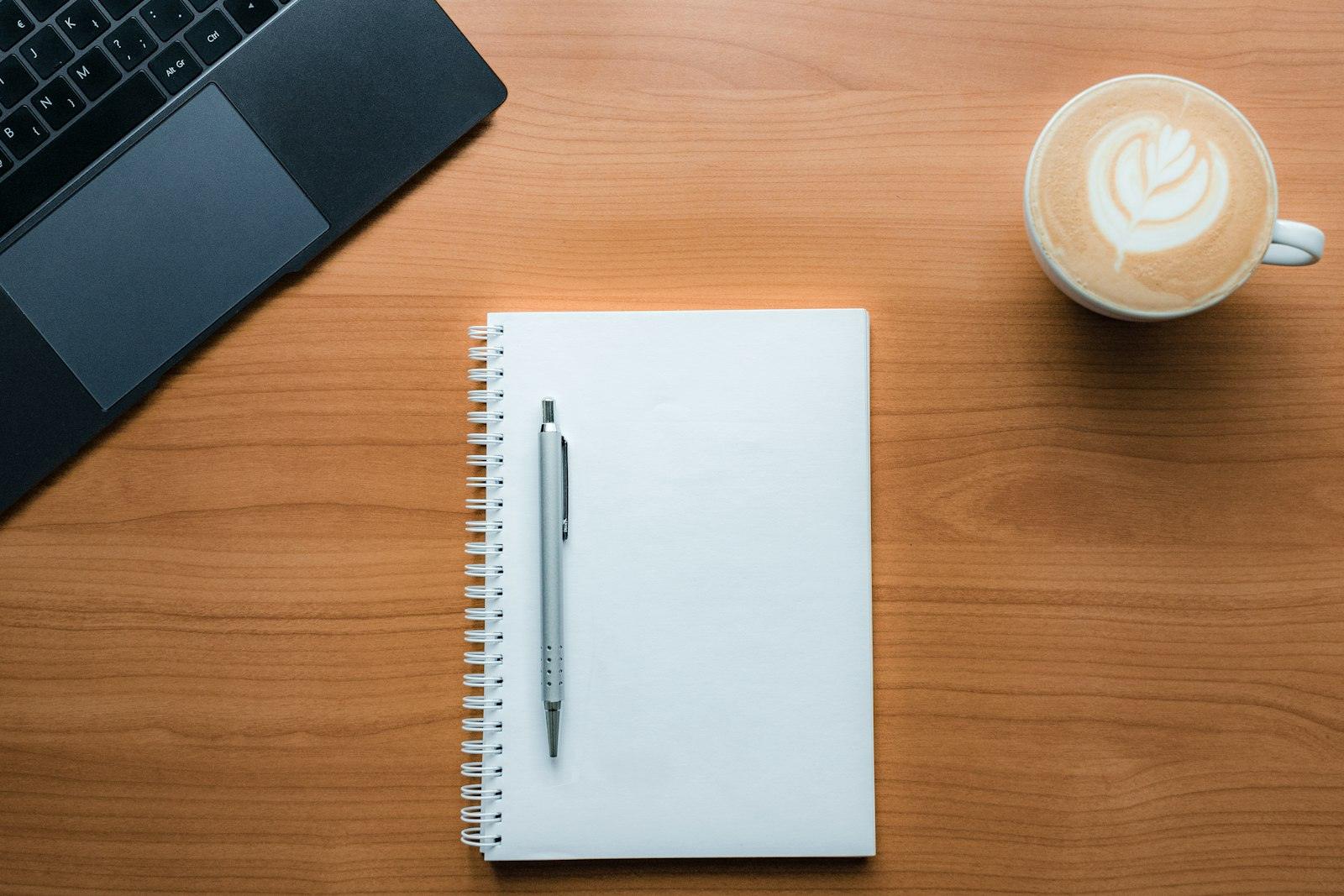 Anteckningsblock med penna, en kaffe latte och en bärbar dator på ett träbord